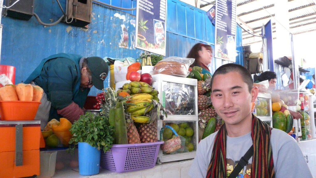 My partner sitting at a juice stand at San Pedro Market in Cusco. There are several fruits on the counter, like pineapples and bananas. There's an old lady behind the counter, who owns the stand.