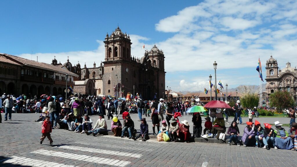 The main square of Cusco, with the Cathedral in the background. There are several people sitting on the sidewalk.