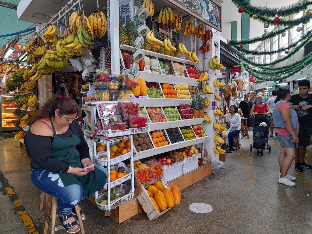 A woman sitting next to her fruit stand in a market in Lima. People walking around the market.