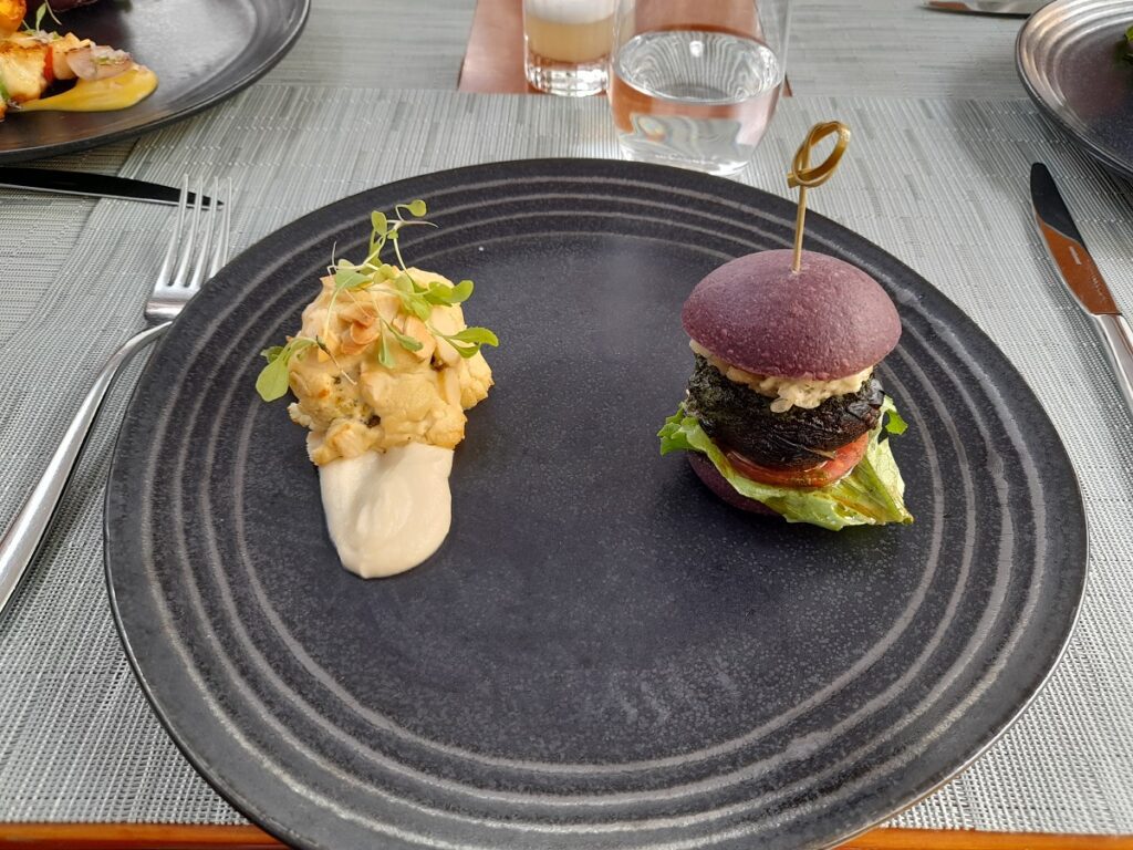 Gourmet vegan portobello mushroom burger served on a unique purple bun, stacked with fresh lettuce and a savory sauce, presented next to a delightful cauliflower creation with a smooth puree, garnished with microgreens, on an elegant dark plate at a fine dining establishment in Lima.