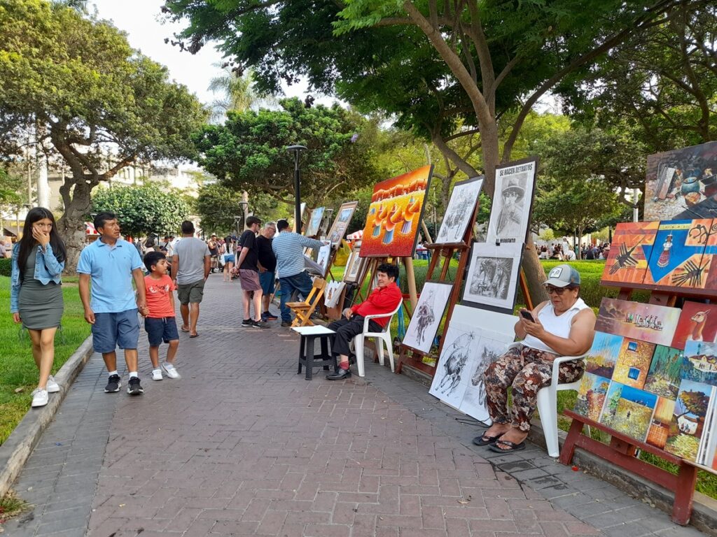 Painters selling artwork at Parque Kennedy, and people walking around, all wearing summer clothes.