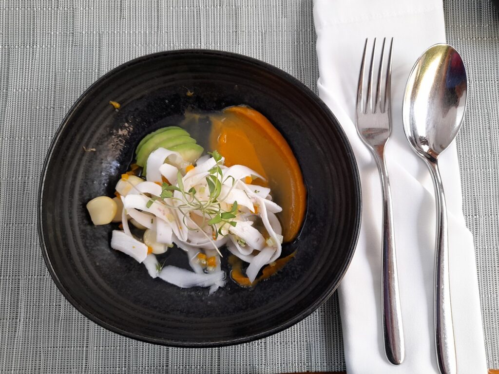 Artfully plated coconut ceviche at Limana restaurant in Lima, featuring tender strips of coconut in a tangy citrus sauce with slices of avocado, corn kernels, and microgreens, all served in a dark, stylish bowl for a refreshing vegan dining experience