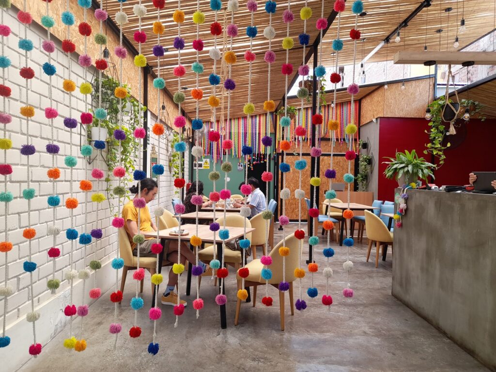 right and inviting vegan gastronomy restaurant in Lima, Peru, with colorful pompoms hanging from the ceiling, creating a cheerful atmosphere. Diners enjoy their meals in a modern, well-lit space with an open roof, lush greenery, and a mix of cozy seating arrangements.