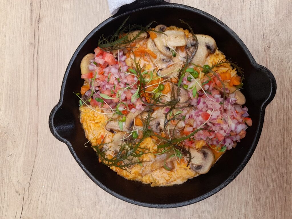 A delectable serving of vegan 'Risotto Marino' in a cast iron skillet, topped with a medley of sautéed mushrooms, fresh diced onion and tomato salsa, and delicate microgreens, presented on a wooden table at Gastronomia Vegana in Lima, Peru, offering a comforting and hearty plant-based meal option