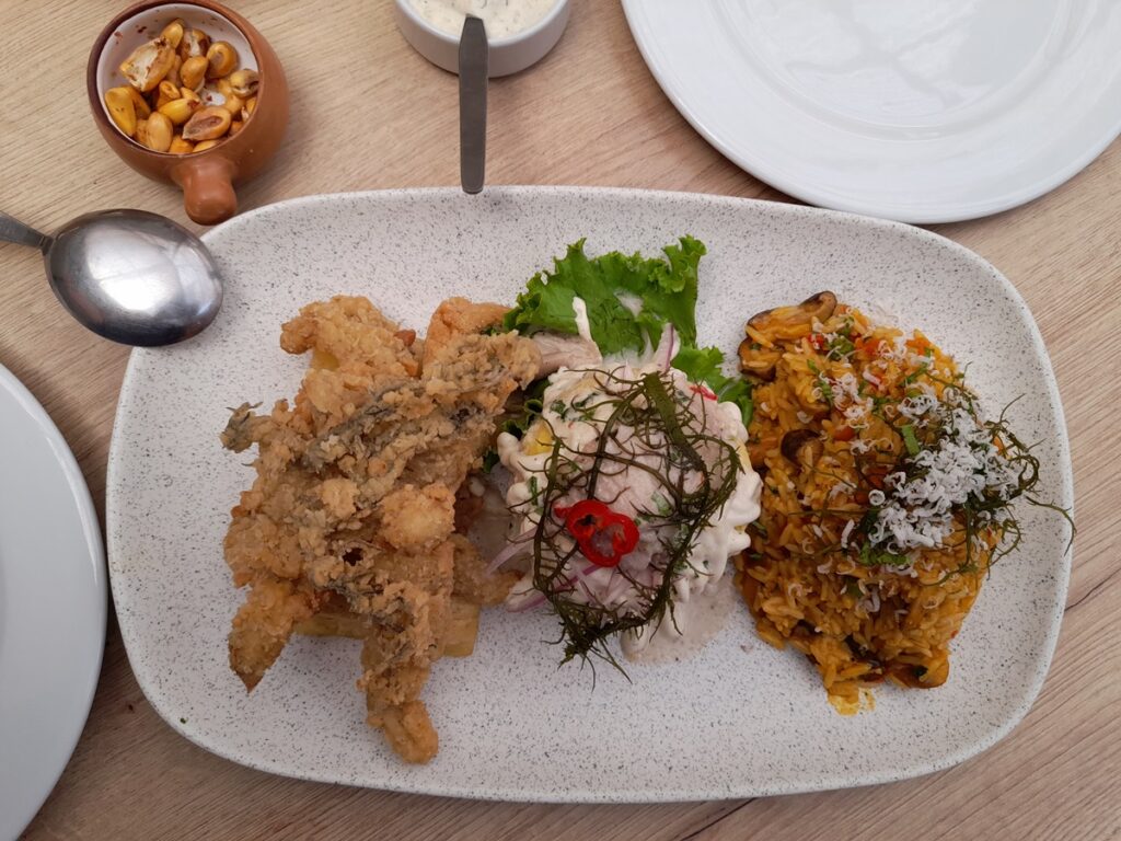 "A delightful Trio Marino platter at Gastronomia Vegana in Lima, offering a taste of the sea with vegan options: crispy mushroom chicharron, a creamy causa topped with chickpea tartar, and a flavorful risotto marino, all beautifully arranged on an oval plate, served with a side of toasted corn kernels.